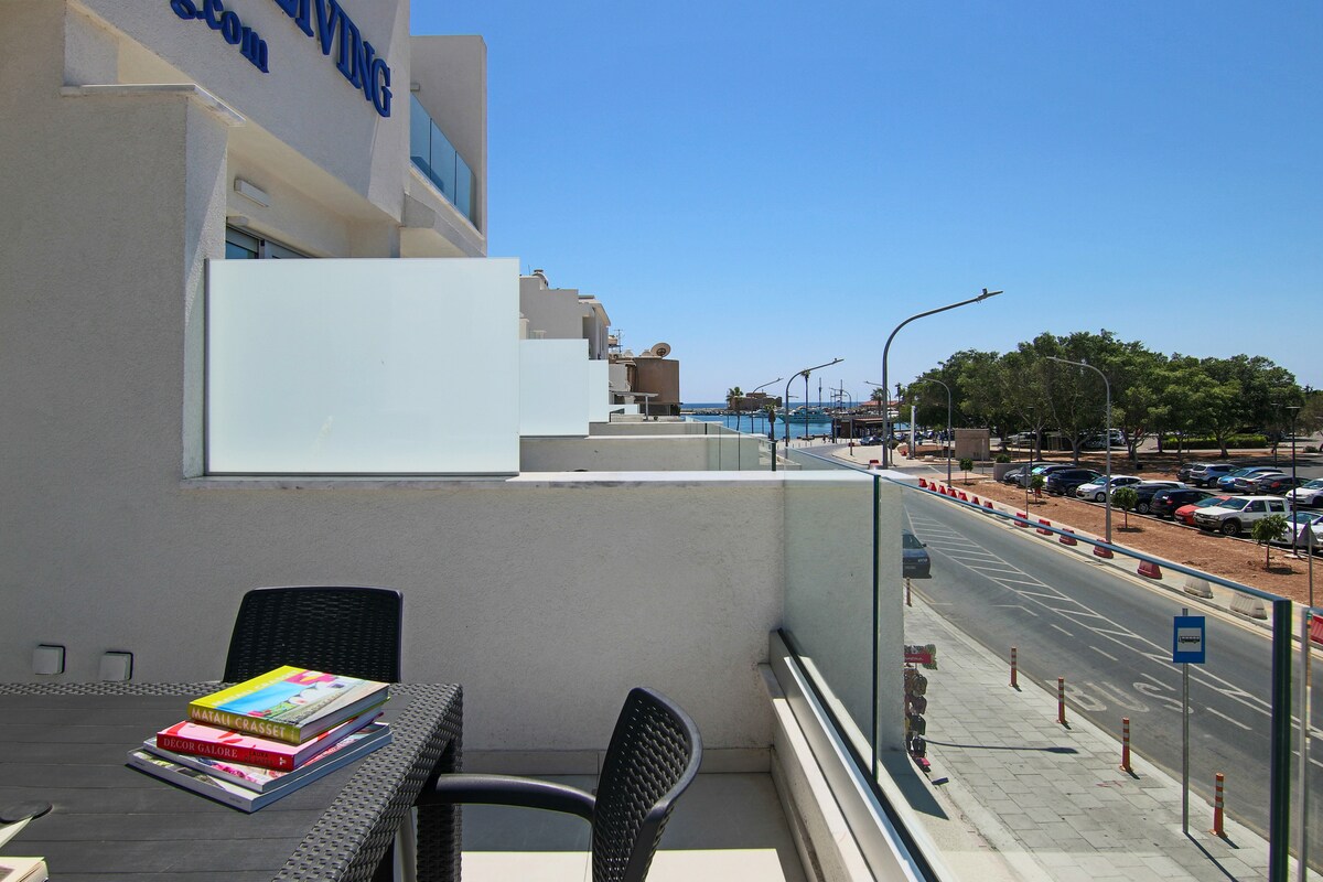 A balcony space is presented with a small table accompanied by two black chairs. A stack of colorful books rests on the table. In the background, the sea is visible beyond a clear glass railing, with trees and a road lined with parked cars.