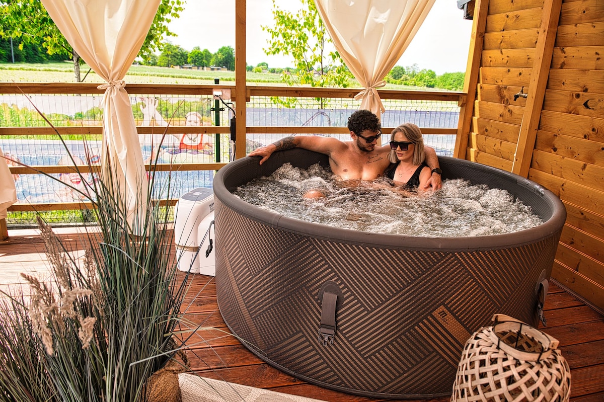 A circular, inflatable jacuzzi is filled with water, with two occupants enjoying the relaxing environment. Surrounding greenery is visible, framed by wooden decking and draped curtains that add a hint of privacy.