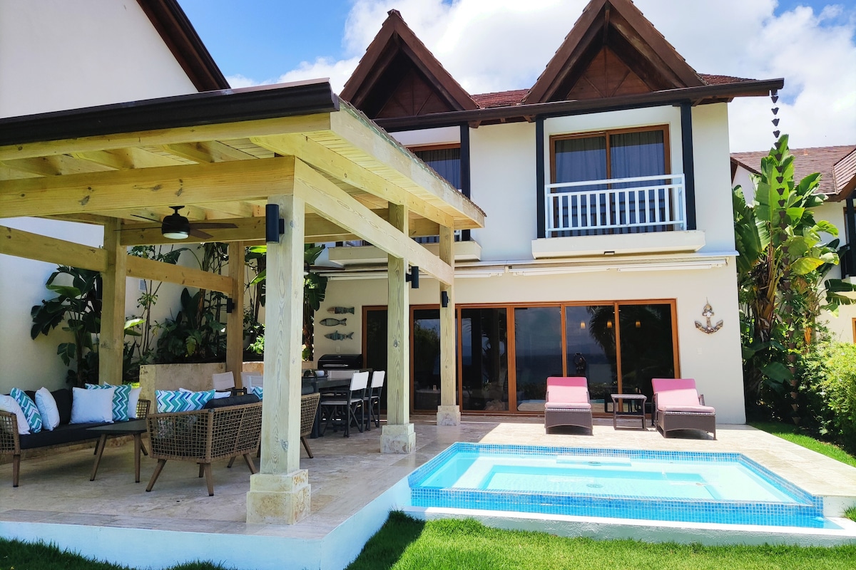 A charming outdoor patio area features a covered dining space with wicker furniture and colorful cushions. Two lounge chairs are positioned beside a small private pool, surrounded by lush greenery. The two-story villa stands in the background with large windows offering views of the surroundings.
