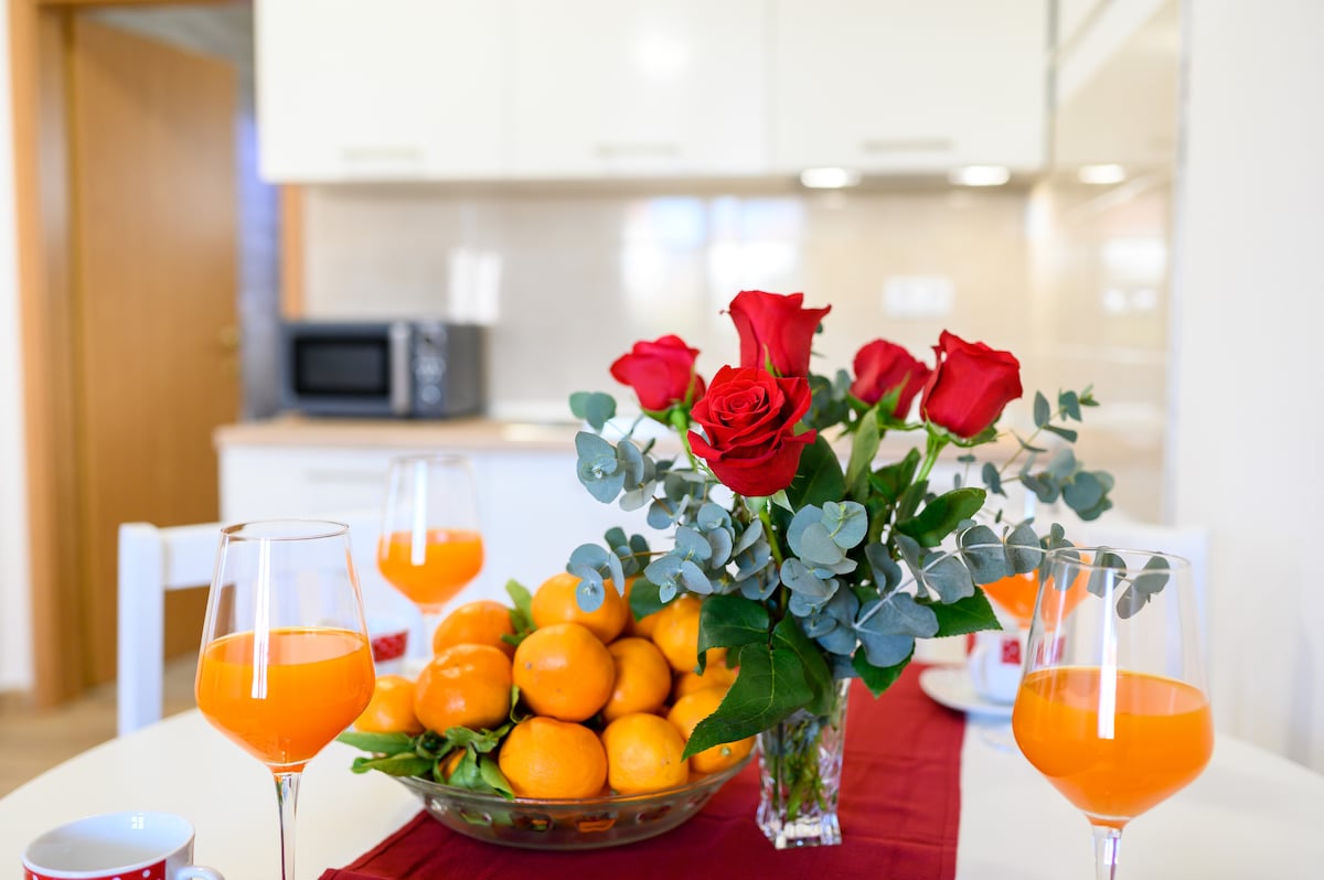 A dining table is set with a bouquet of red roses and a bowl of oranges. Two glasses filled with orange juice sit next to the bowl. The background features a modern kitchen with light cabinetry and a microwave.