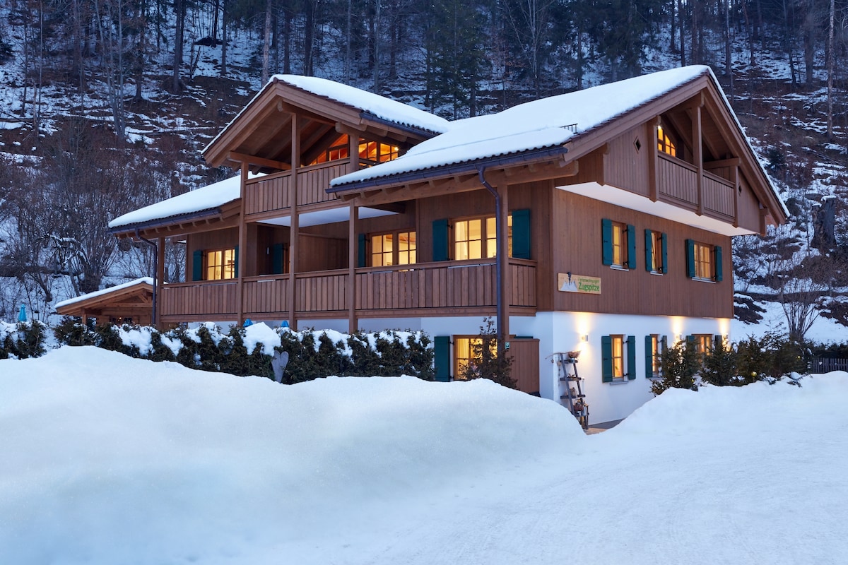 A chalet-style building with wooden balconies and multiple warm-lit windows is surrounded by snow-covered terrain. The structure features a welcoming design, showcasing natural materials and the charm of mountain architecture.
