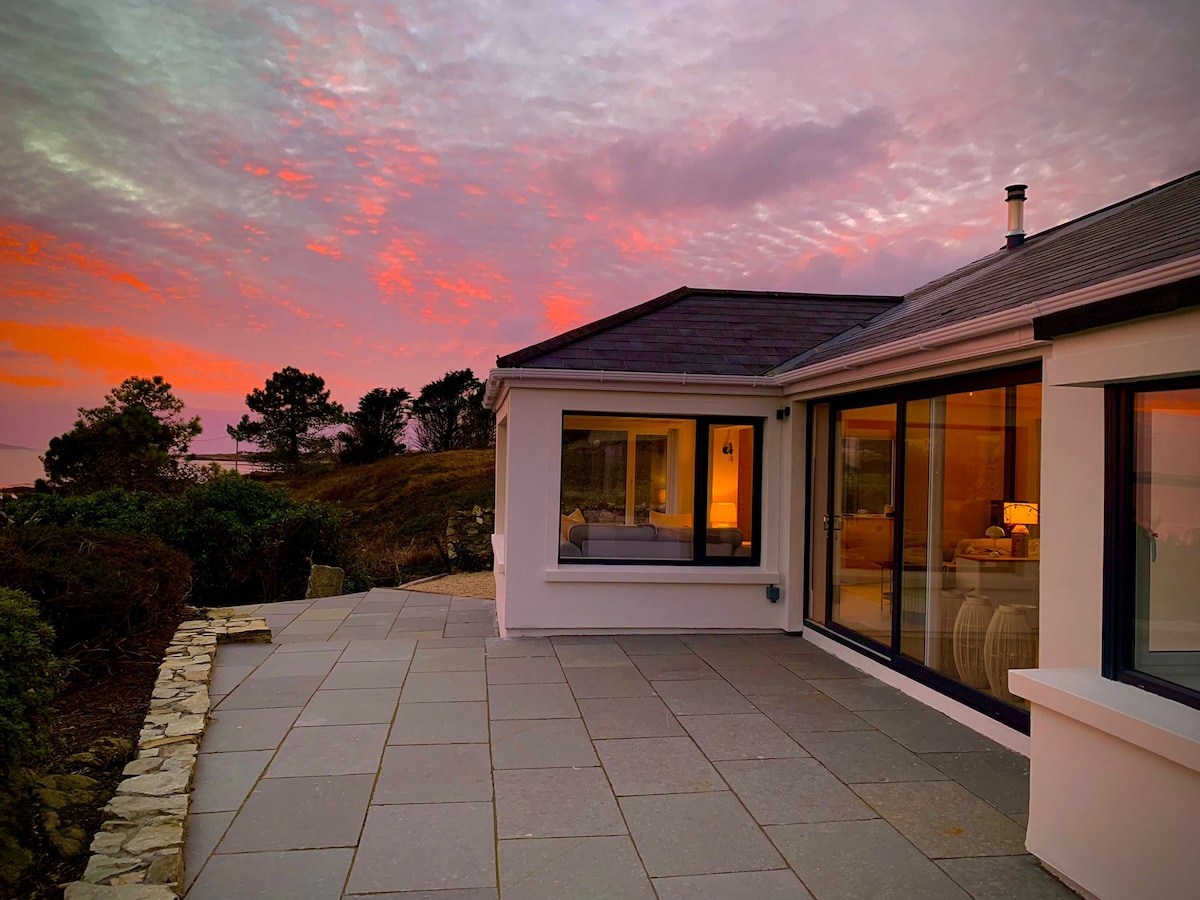 The exterior of a newly renovated home is showcased against a vibrant sunset sky, with large glass windows reflecting warm tones. A spacious stone patio extends from the house, bordered by lush greenery and offering a seamless connection to the serene landscape.