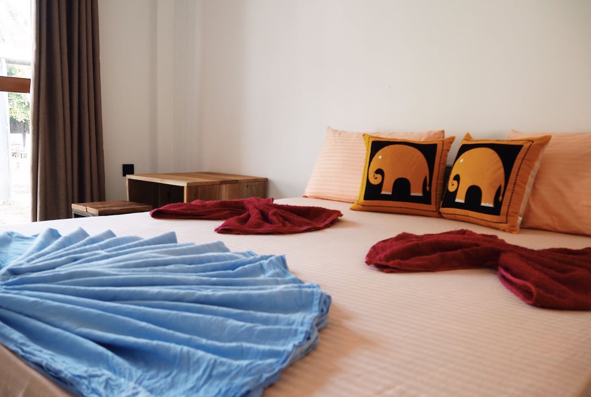 A spacious bed is adorned with two decorative pillows featuring elephant designs. Light blankets in blue and maroon are artistically arranged across the bedding. Natural light filters in through a window, complemented by textured curtains and a wooden bedside table.