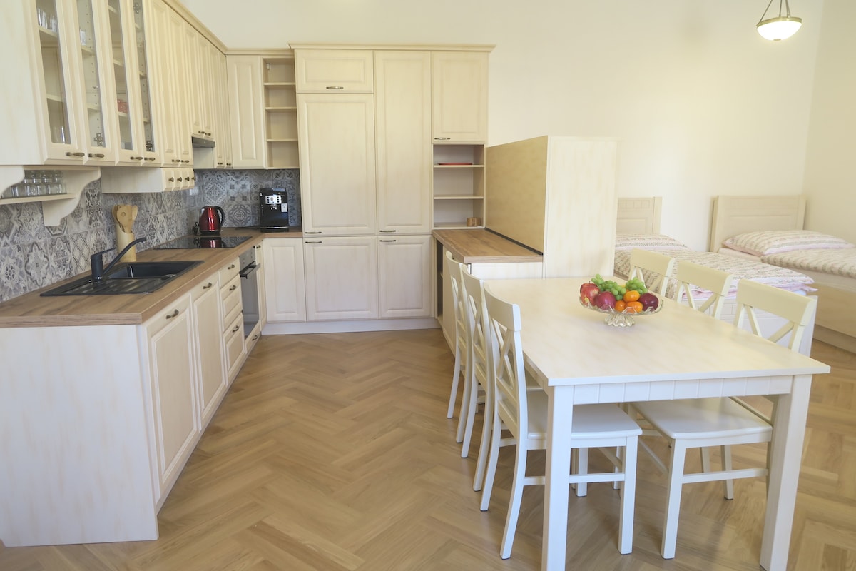 A functional kitchen area features light wood cabinetry and a dining table set for six. The kitchen includes modern appliances, with a fruit bowl at the center of the table. Two single beds are positioned in the background, complementing the open layout.