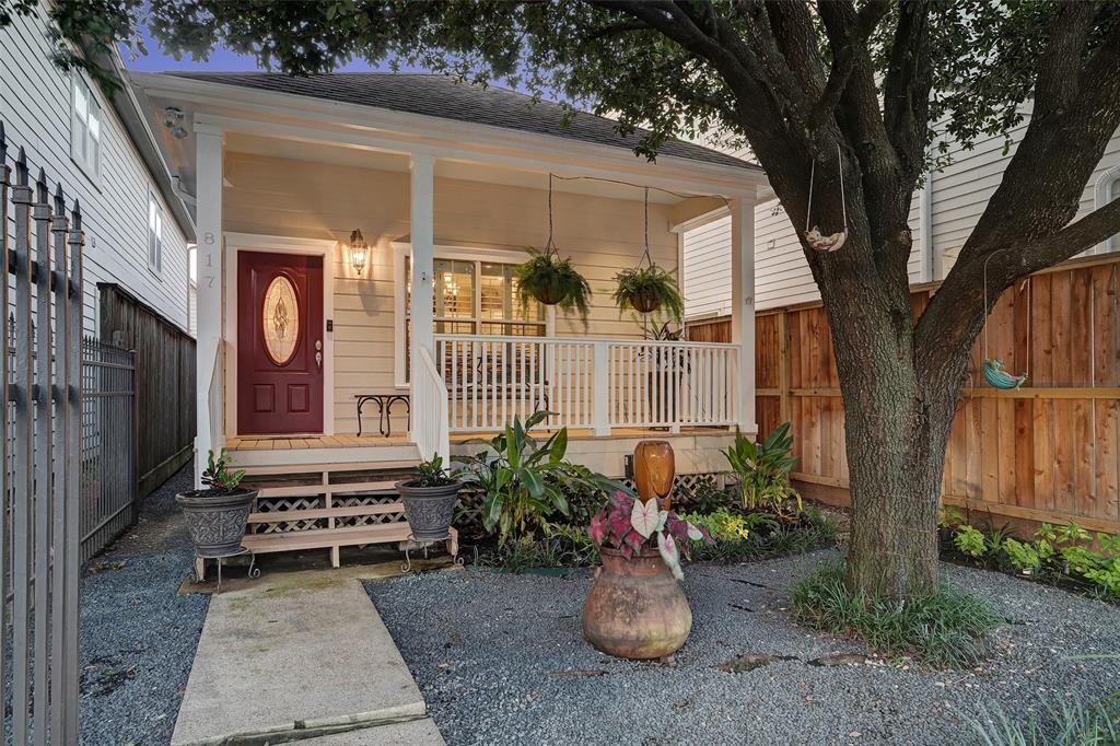 The entrance features a welcoming porch with white railings and potted plants, set beneath a large tree. A front door in a rich burgundy hue is highlighted, and an earthy ceramic pot is positioned on the gravel walkway that leads to the entrance.