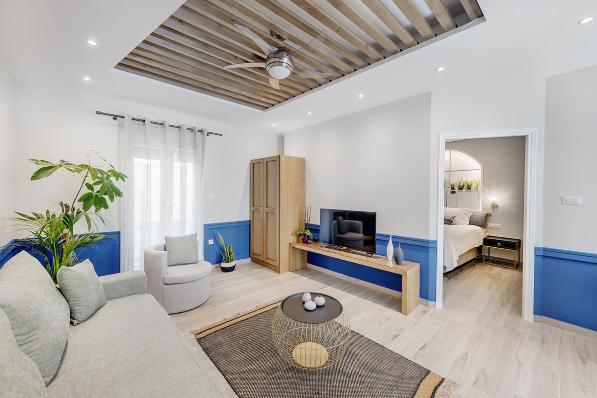 A modern living area is highlighted by a comfortable gray sofa and a round coffee table, with a ceiling fan above for airflow. A sleek television rests on a wooden console against a light wall. Decorative plants enhance the space, and a doorway leads to the bedroom.