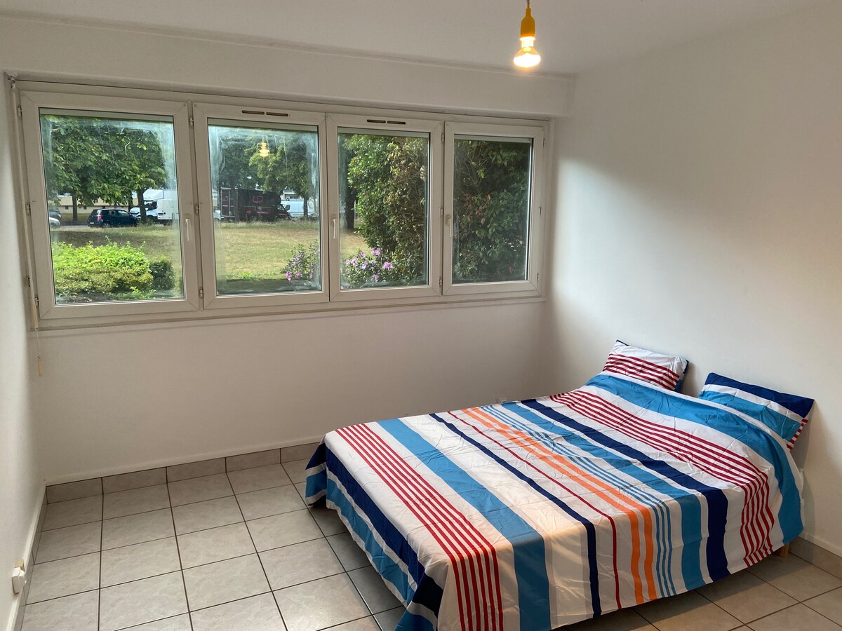A bright sleeping area features a spacious bed dressed in a striped blue and white bedding set. Large windows provide natural light and offer views of greenery outside. The tiled floor adds a clean and modern feel to the room.