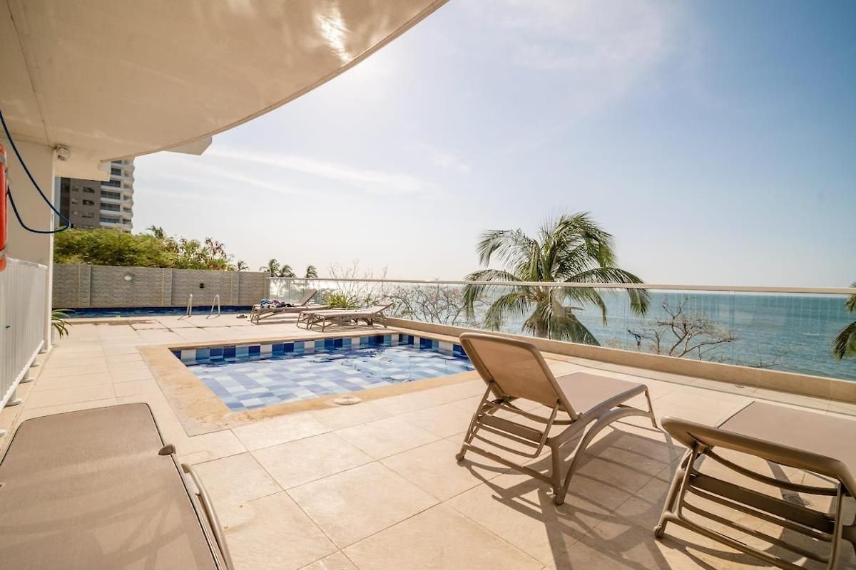 A terrace area is shown with two lounge chairs positioned beside a small pool. Clear views of the ocean are visible beyond the railing, with palm trees framing the scene. The tiled patio is sunlit, creating a calm and inviting outdoor space.