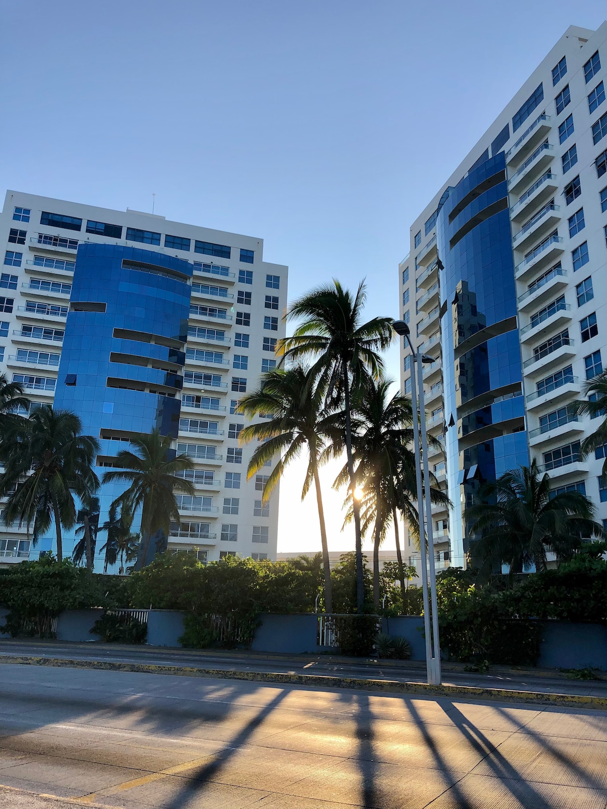 Two modern buildings with reflective glass exteriors are framed by palm trees. The sunlight creates a soft glow behind the structures, casting long shadows on the street below. The overall setting conveys an inviting urban environment near the beach.