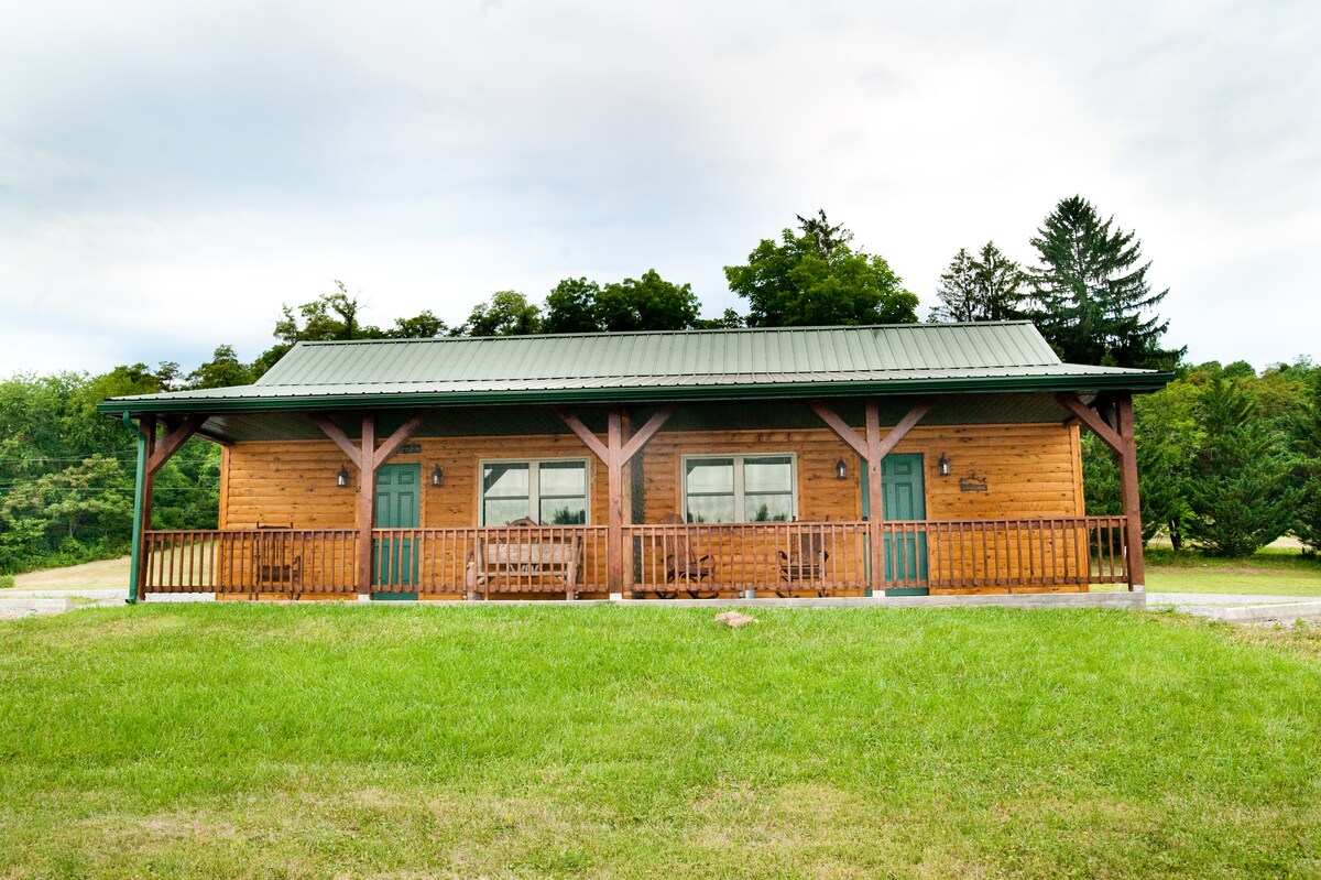 A log cabin structure featuring a green metal roof and a welcoming front porch with wooden seating. The exterior is surrounded by expansive green grass and tall trees, providing a serene setting. Large windows allow natural light to illuminate the interior.