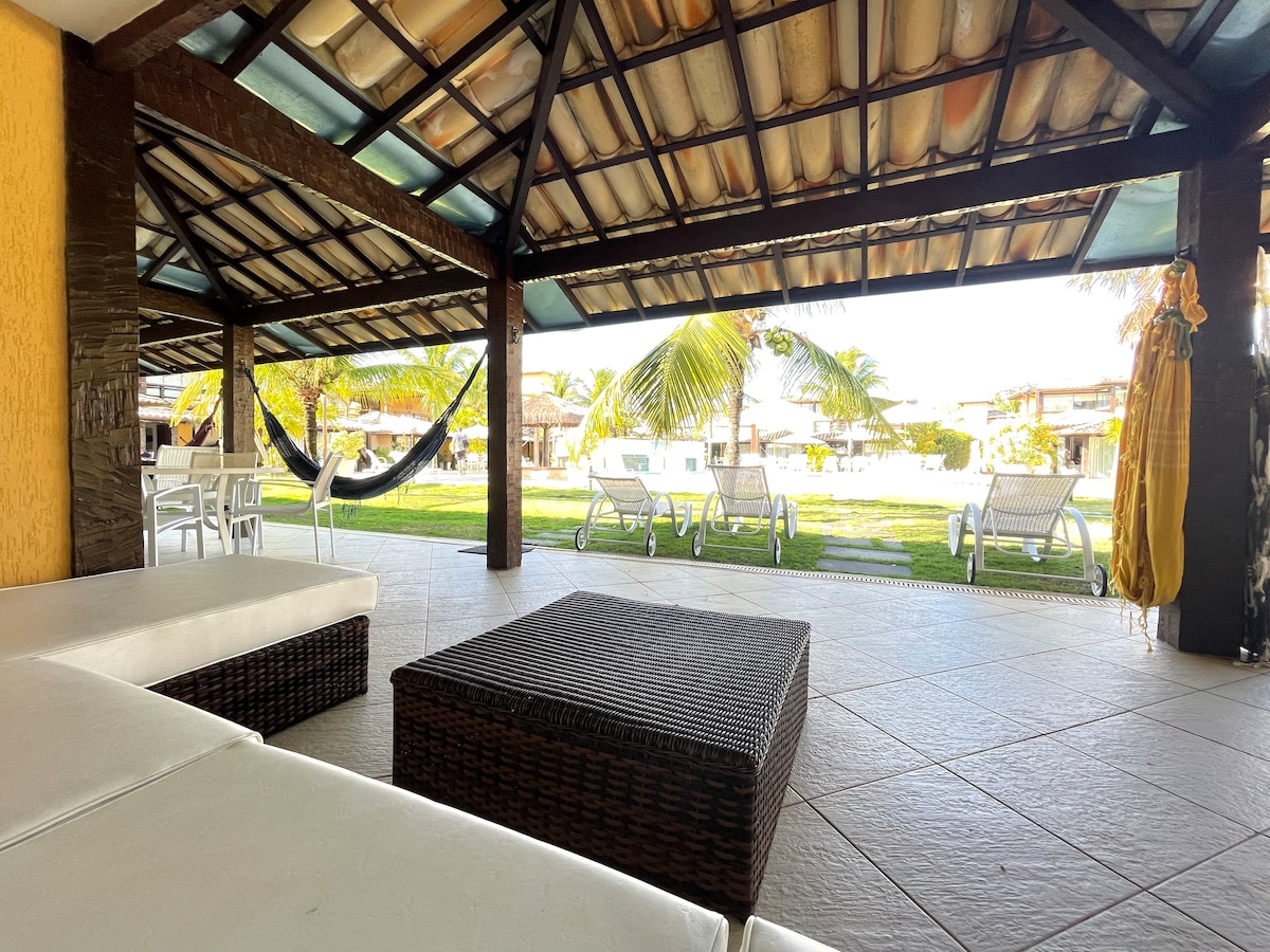 A shaded outdoor lounge area is visible, featuring a low wicker table and seating arrangements. Behind, sun loungers and a hammock are positioned on a grassy expanse, with palm trees swaying gently in the breeze. The structure is supported by wooden beams and a tiled roof.