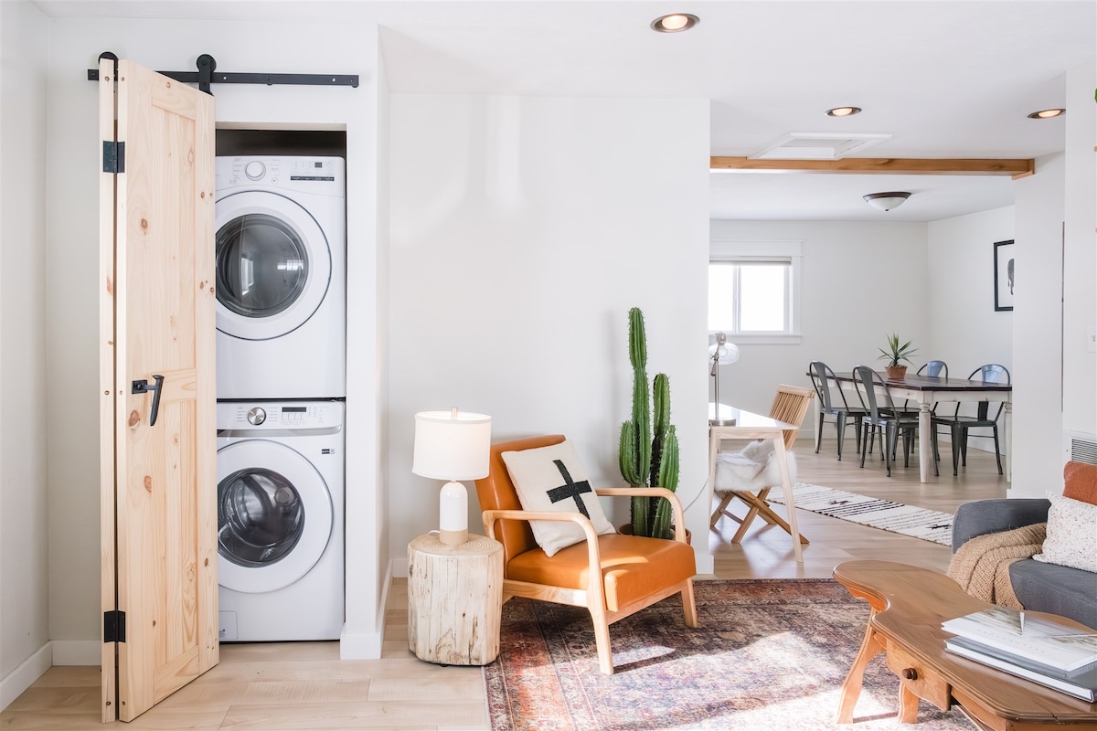 A laundry area is integrated into the living space, featuring stacked washer and dryer units behind a wooden sliding door. Nearby, a comfortable armchair sits beside a table lamp, with a cactus adding a touch of greenery, and a dining area is visible in the background.
