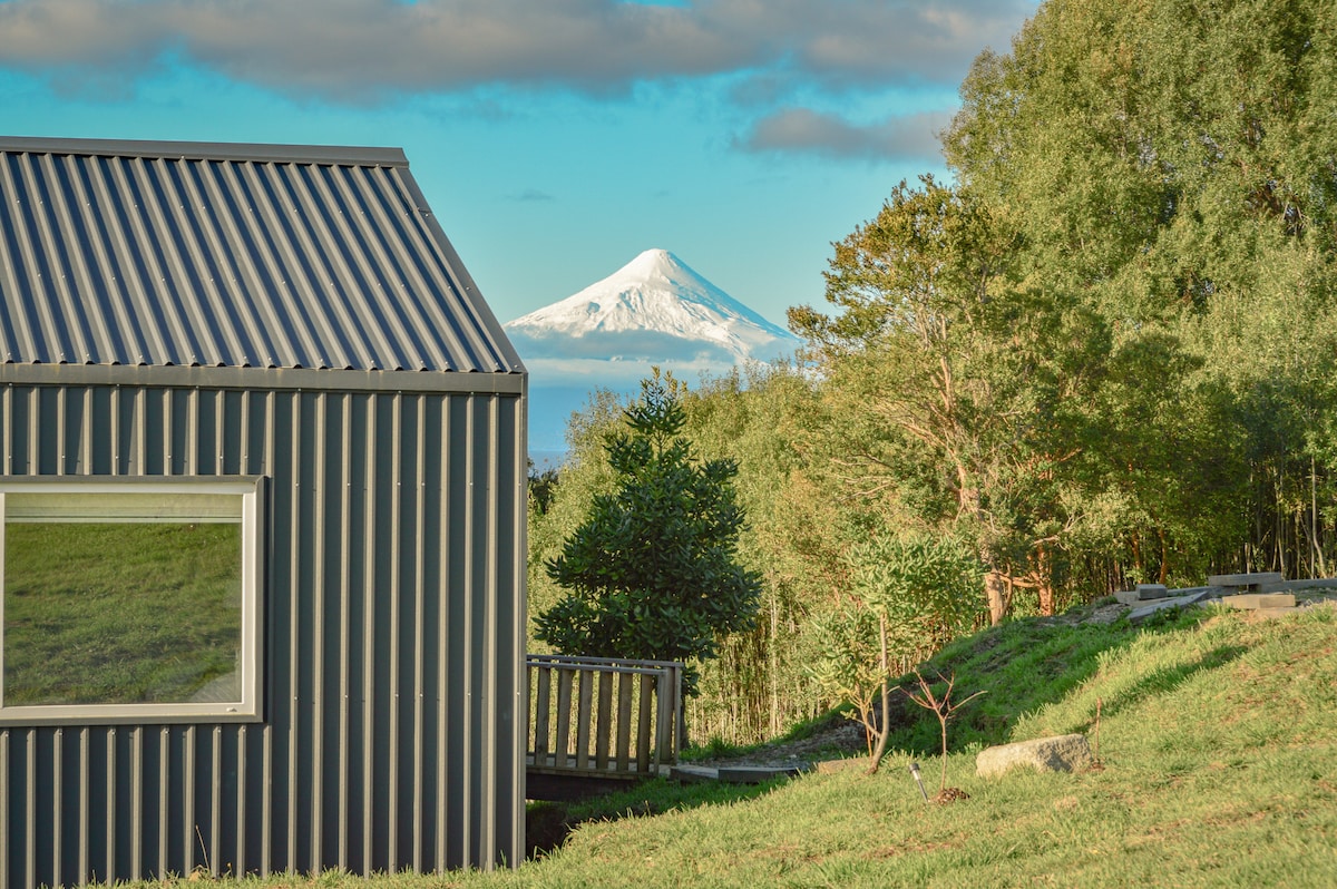 A modern cabin with a sleek metal exterior is framed by lush greenery. The snow-capped peak of a volcano is visible in the background, contrasting against the blue sky. A wooden pathway leads to the cabin, enhancing the connection with nature.