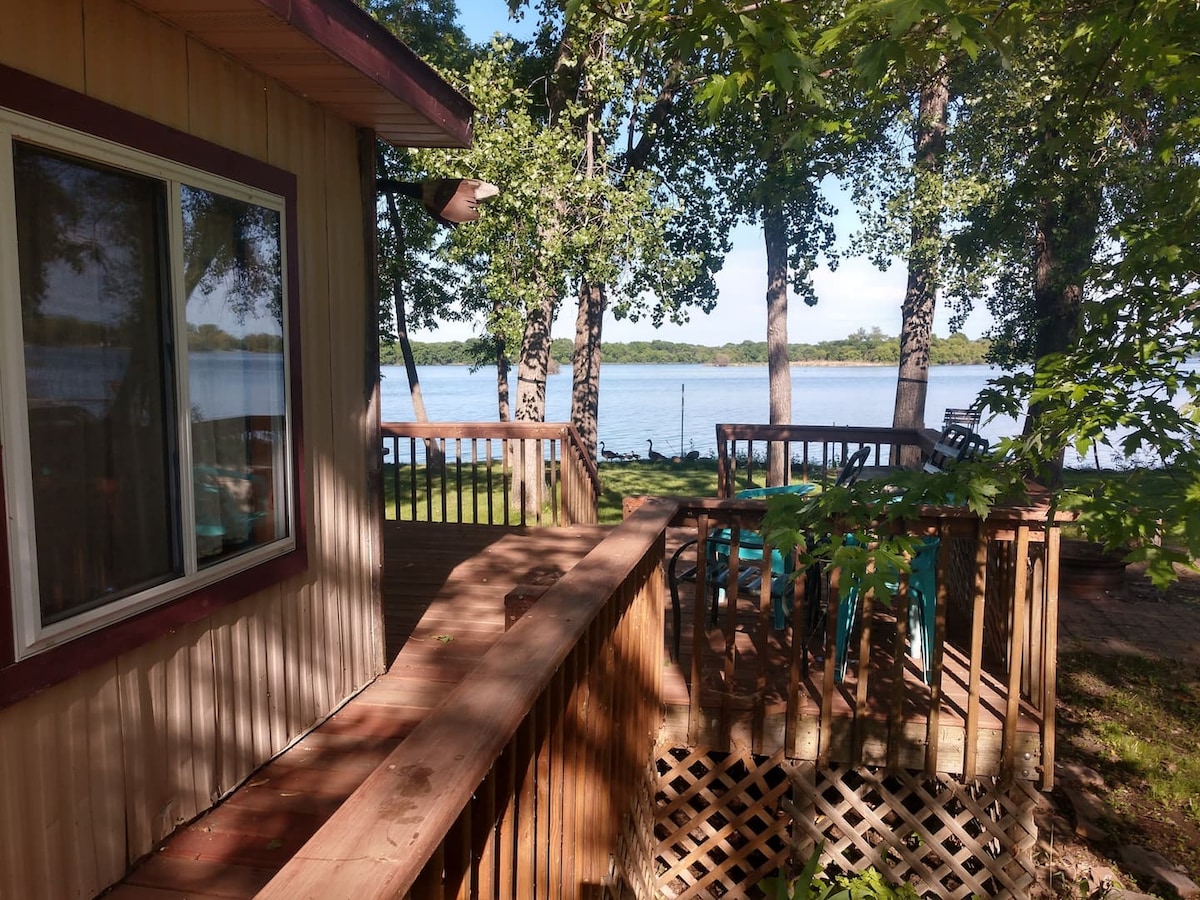 A cabin exterior is framed by lush greenery, overlooking a calm body of water. A wooden deck, equipped with patio furniture, extends towards the shoreline, providing a serene area for relaxation. Large windows reflect natural light, enhancing the inviting atmosphere.