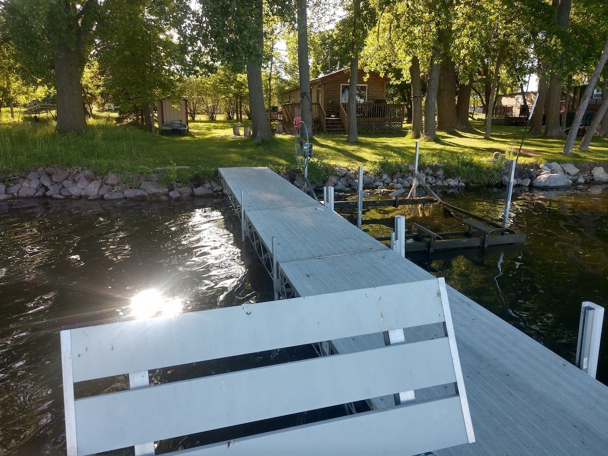 A wooden dock stretches out into the water, bordered by lush green trees. The distant cabin is visible, nestled among the foliage, with a shaded area created by overhanging branches. The sunlight glimmers off the water, providing a peaceful atmosphere.