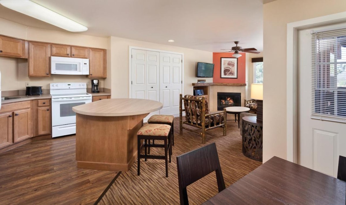The open-plan living area features a well-equipped kitchen with wooden cabinetry and modern appliances. A dining table and chairs are arranged near the kitchen. A cozy seating area includes decorative chairs facing a gas fireplace, while a wall-mounted TV completes the inviting space.