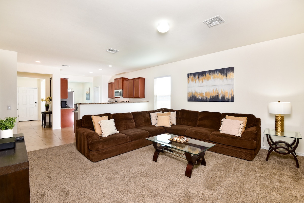A spacious living area features a large brown sectional sofa arranged around a glass coffee table. Soft beige carpeting covers the floor, and natural light enters through the windows. A modern piece of wall art adds a touch of color, complementing the warm tones of the room.