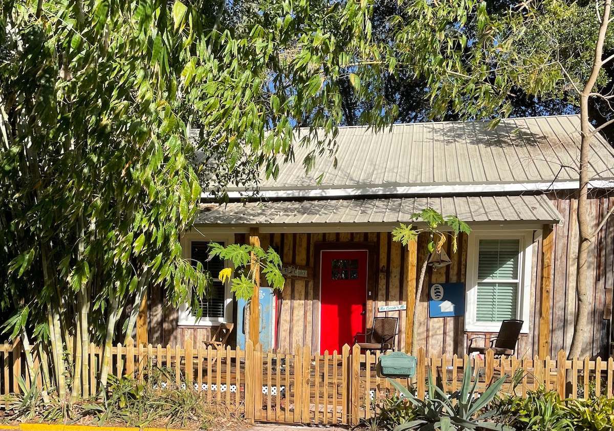 The cottage exterior is adorned with classic wooden siding and features a vibrant red door. Flanking the entrance are two cozy chairs, set against a backdrop of lush greenery and a decorative fence, enhancing the inviting appeal of the space.