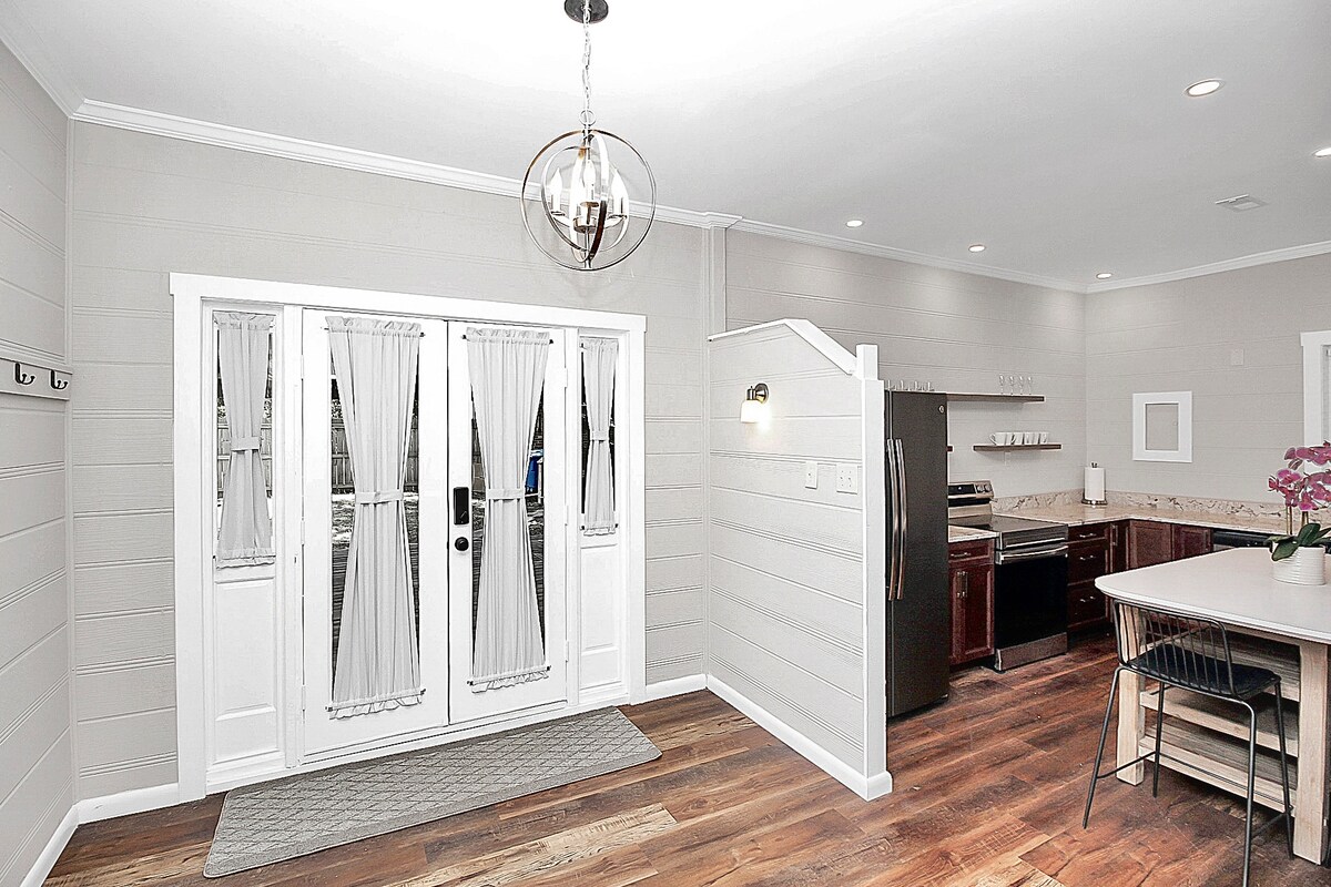 The entrance features double doors with sheer curtains, granting a soft light. A welcoming area is surrounded by hardwood flooring and neutral-colored walls, complemented by a modern light fixture above. A kitchen area is visible, with dark cabinetry and stainless steel appliances providing a contrast to the inviting space.