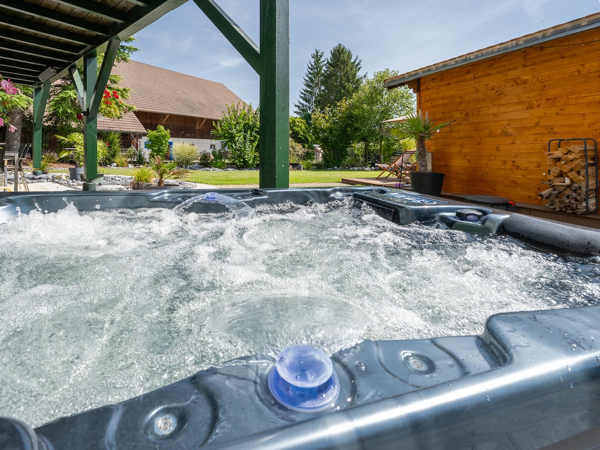 A hot tub bubbles under a shaded pergola, surrounded by greenery and vibrant flowers. Soft sunlight filters through the space, highlighting the wood-paneled shed in the background. The peaceful outdoor setting invites relaxation and moments of tranquility.