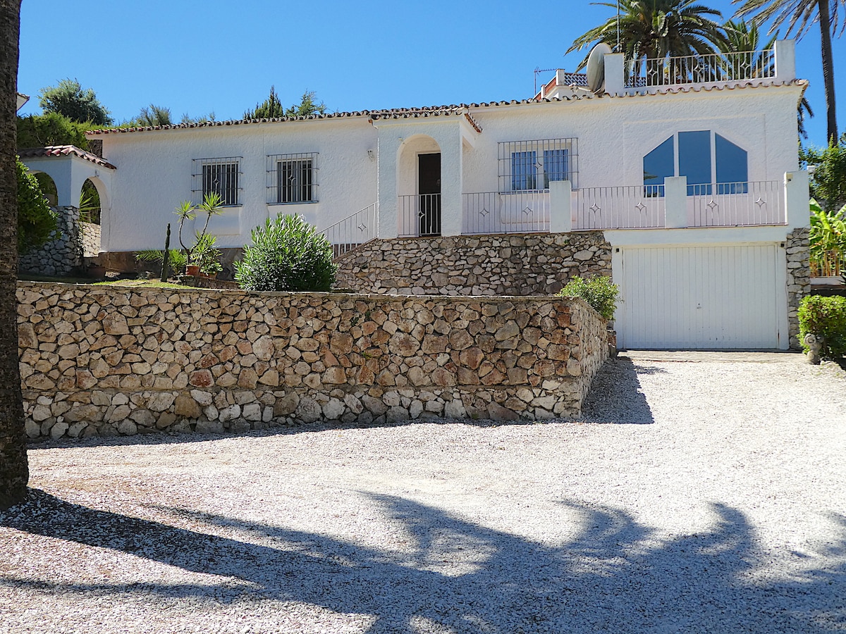 A light-colored, two-story vacation house features a prominent balcony and large windows. The front of the house is bordered by a natural stone wall, with a gravel driveway leading to a garage. Lush plants and palm trees are visible in the sunny landscaped area.