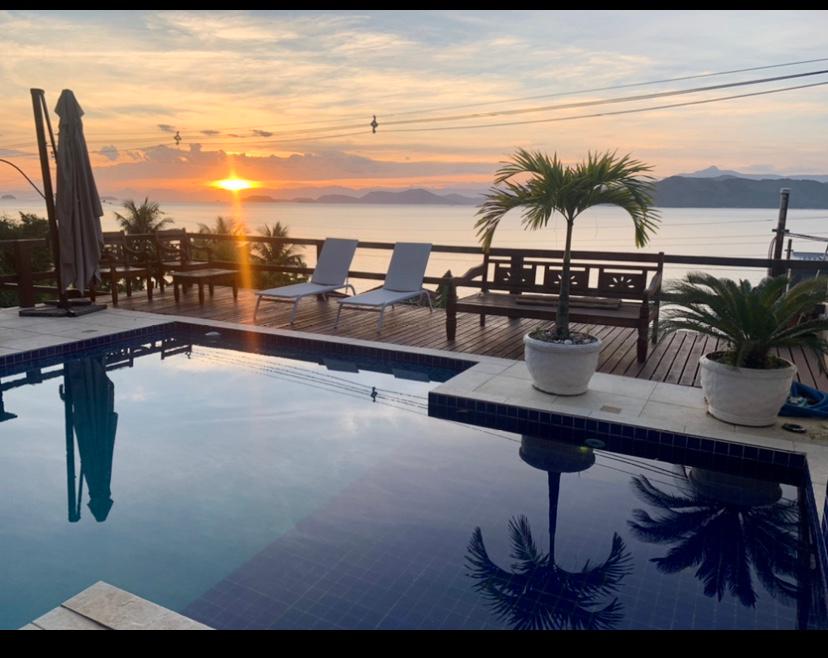 A tranquil pool area features a clear blue pool reflecting the sunset, framed by wooden decking. Lounge chairs are positioned near the edge, while palm trees and potted plants add greenery. The serene view includes distant mountains and an expansive sky filled with warm colors.