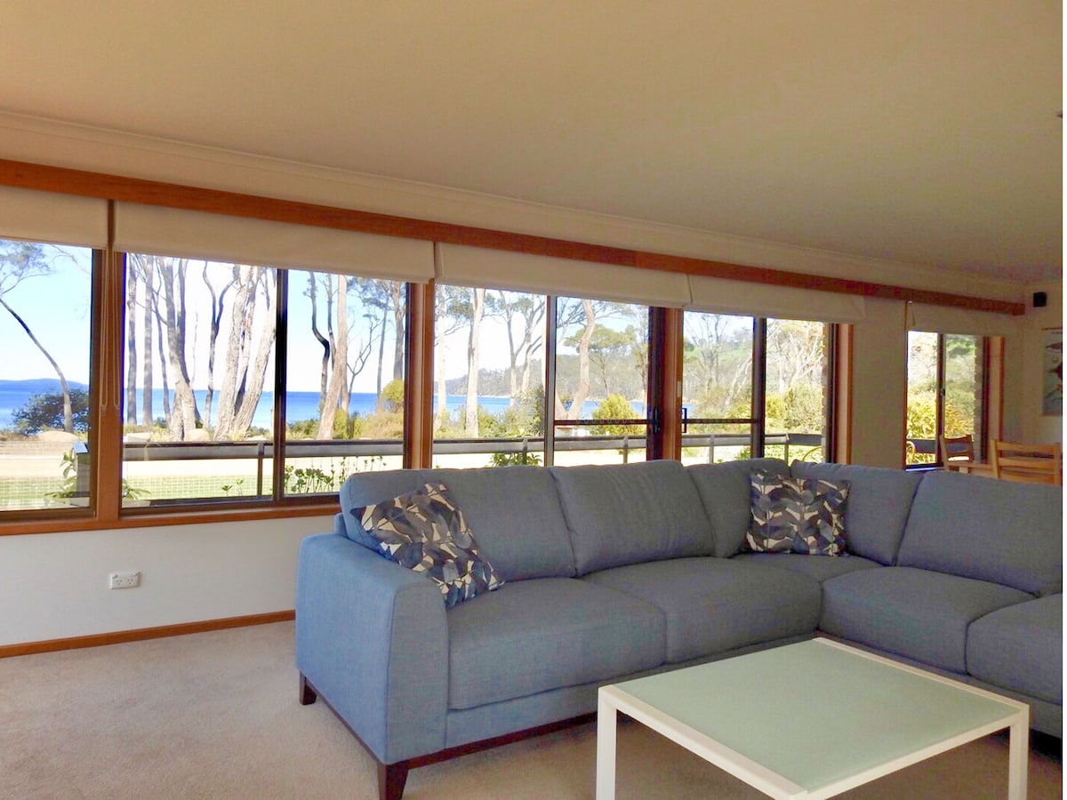 A spacious living area features a large blue sectional couch adorned with decorative pillows. Expansive windows provide a view of the serene waters of Roaring Bay, allowing natural light to illuminate the interior.