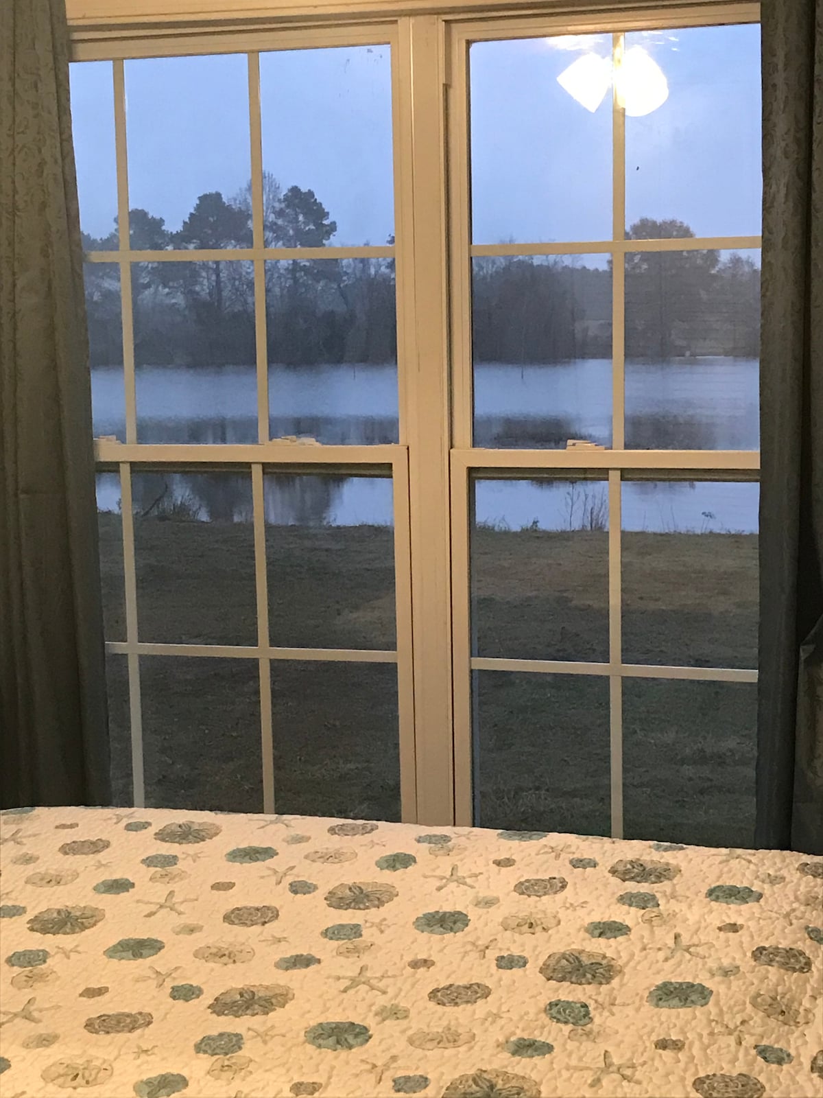 A large window frames a view of a serene pond surrounded by grass and trees, with the soft light of dusk filtering through. The window features multiple panes, and a patterned bedspread with subtle colors is visible in the foreground.