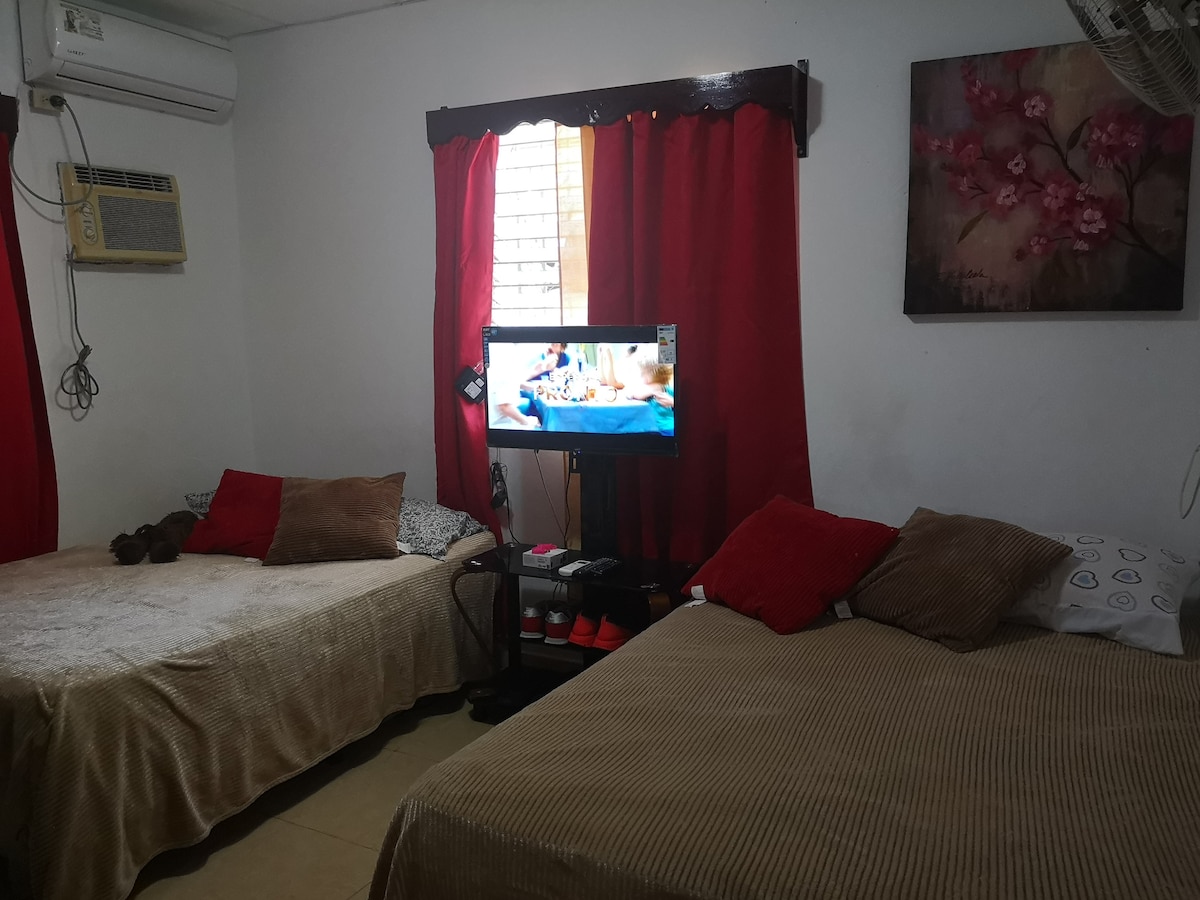 A room features two spacious beds, each adorned with textured bedding and decorative pillows. A unit air conditioner is mounted on the wall, and a television is positioned on a small stand, providing entertainment. Natural light enters through a window covered by red curtains.