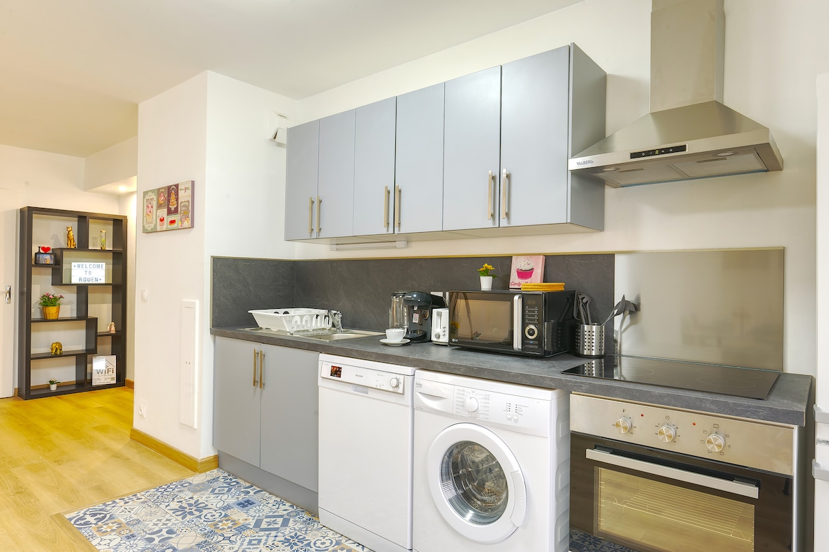 The kitchen area features modern cabinetry in soft gray, complete with a variety of appliances, including a stove, microwave, and dishwasher. A washing machine is also present. The countertop is complemented by a stylish backsplash and organized kitchen essentials, creating an efficient cooking space.