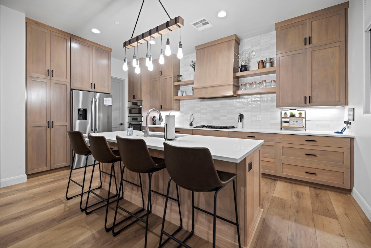 A modern kitchen features wooden cabinetry and a spacious island with seating for four. A stylish pendant light illuminates the area. Stainless steel appliances are integrated, and neutral tiles enhance the overall design. Countertop space is available for meal preparation.