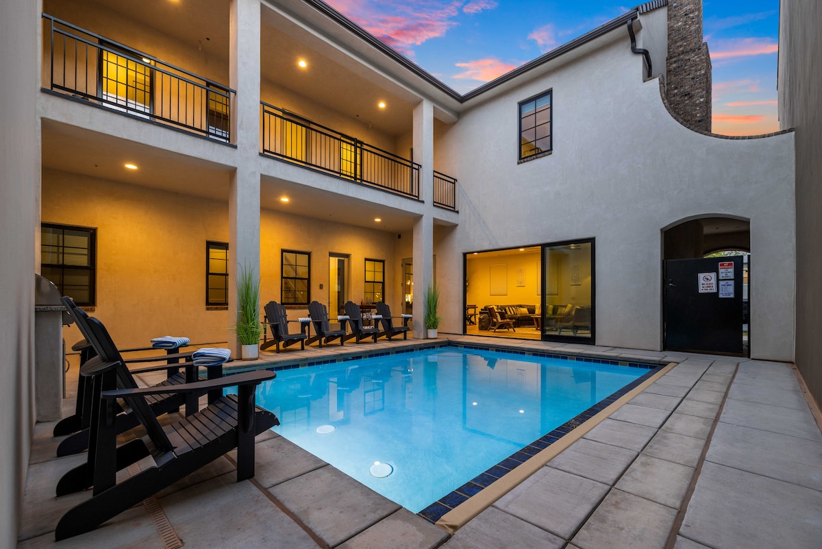 A private pool is surrounded by a tiled patio with several loungers positioned along the edges. Two stories of the property can be seen, with large windows providing views of the indoor spaces. Soft lighting enhances the ambiance as evening approaches.