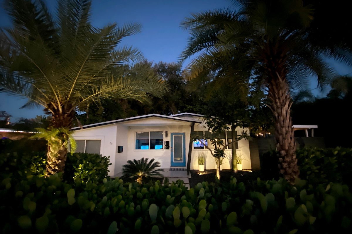 The exterior of the house is illuminated by soft outdoor lighting, showcasing a well-maintained facade and tropical landscaping. Towering palms flank the entrance, while lush greenery provides a natural border around the property, creating a welcoming ambiance as night falls.