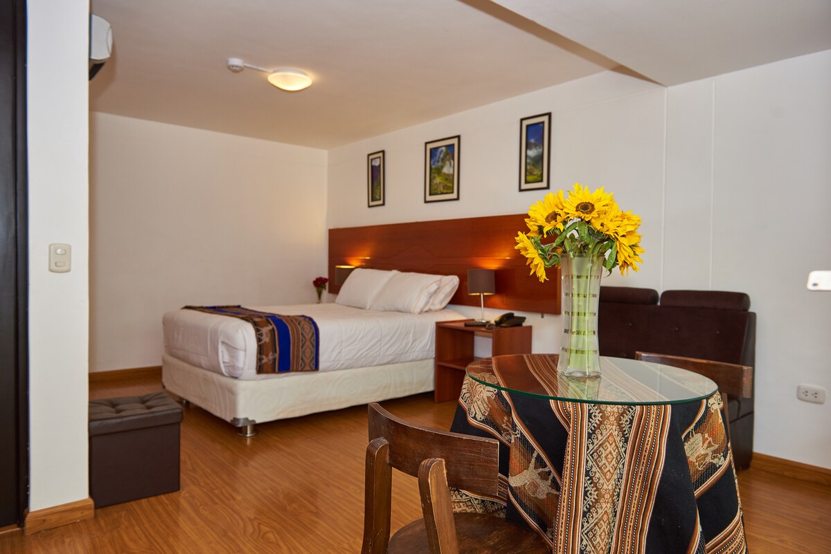 The room features a spacious bed with a wooden headboard, complemented by colorful artwork on the walls. A small round table is set with a floral arrangement, and a cozy chair is positioned nearby. Warm wood flooring enhances the inviting atmosphere.