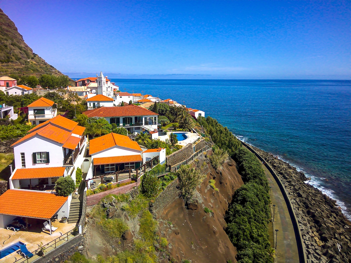 The aerial view showcases the villa's white structure with red-tiled roofs, positioned along the coastline. The blue sea stretches toward the horizon, while the surrounding greenery and neighboring houses create a serene scene. A swimming pool is visible on the villa's lower terrace.