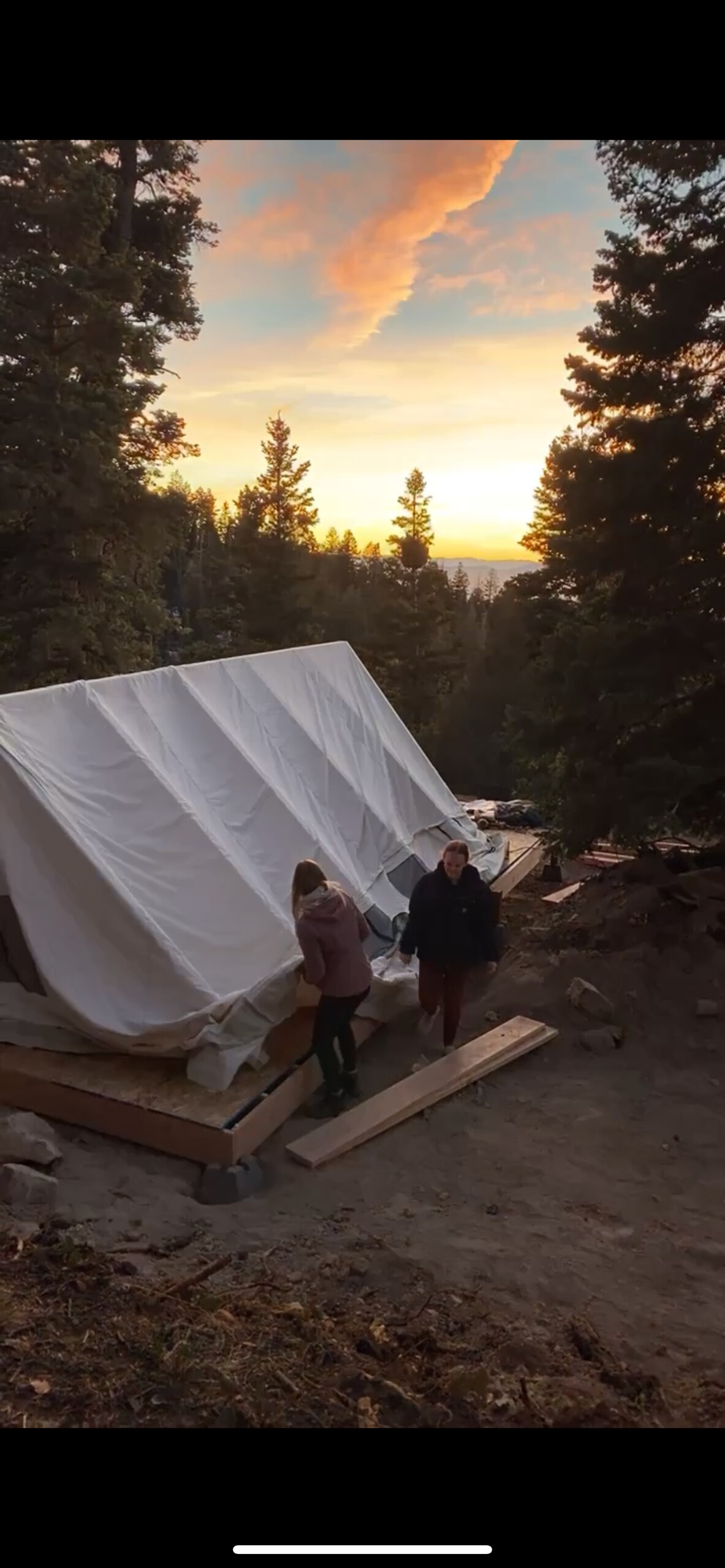 A private tent is set on a wooded lot, surrounded by tall trees. The sunset casts a warm glow, highlighting the natural landscape. Two figures stand near the entrance of the tent, creating a sense of tranquility in a secluded area.