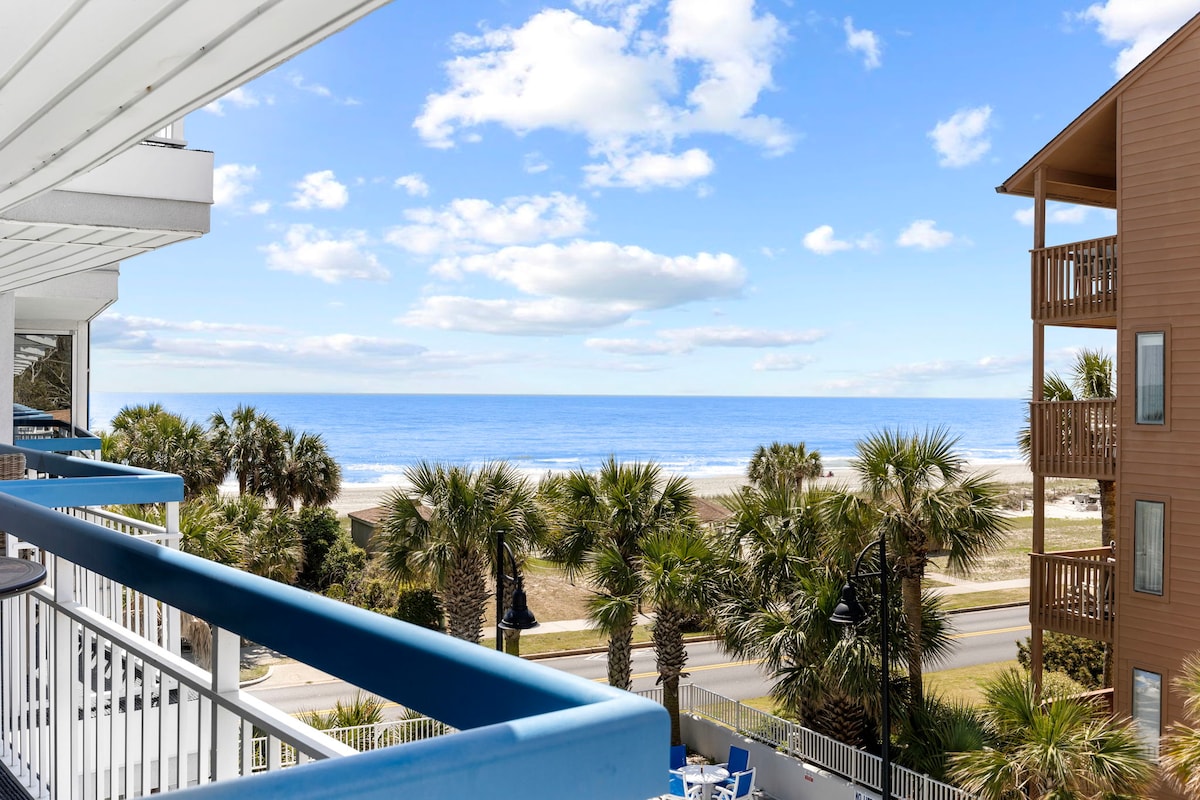 A balcony view showcases the expansive Atlantic Ocean under a clear blue sky, framed by palm trees and a sandy beach. The scene includes a well-maintained pathway leading to the shoreline, with a decorative railing in the foreground.