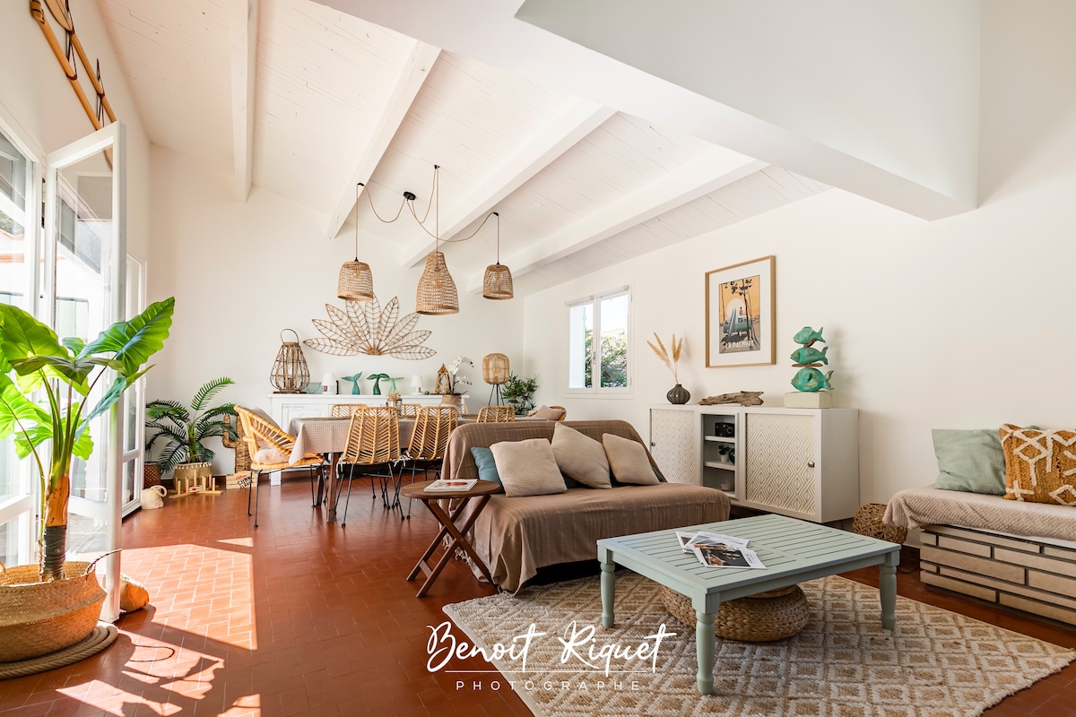 A spacious living area features high ceilings with wooden beams and large windows allowing ample natural light. Comfortable seating arrangements include a sofa and a coffee table. Decorative elements include woven pendant lights and plants, creating a relaxed atmosphere adjacent to the dining space.