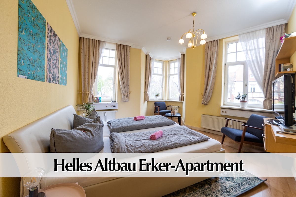 A bright apartment features large windows dressed with soft curtains, allowing natural light to fill the space. Two spacious beds are positioned against a pastel yellow wall, with decorative wall art enhancing the ambiance. A TV sits on a wooden stand, providing entertainment options.