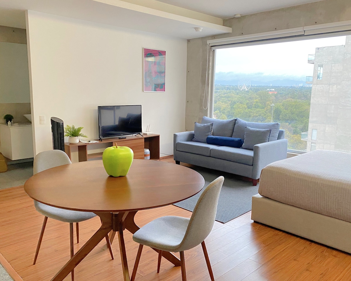 An open-concept living space features a circular dining table with four chairs, a cozy sofa against a large window, and a smart TV on a stand. A green apple sits at the center of the table, and the backdrop showcases views of trees and the skyline.