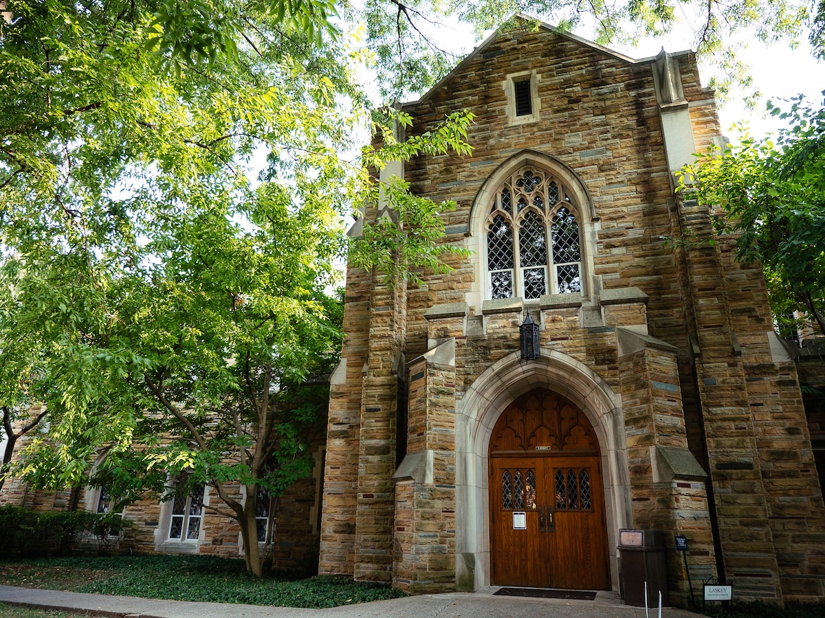 The exterior of a historic building features intricate stonework and gothic-style architecture. Tall arched windows adorned with decorative grills allow natural light to filter through. Lush greenery surrounds the entrance, which is highlighted by a set of large wooden doors beneath a prominent archway.