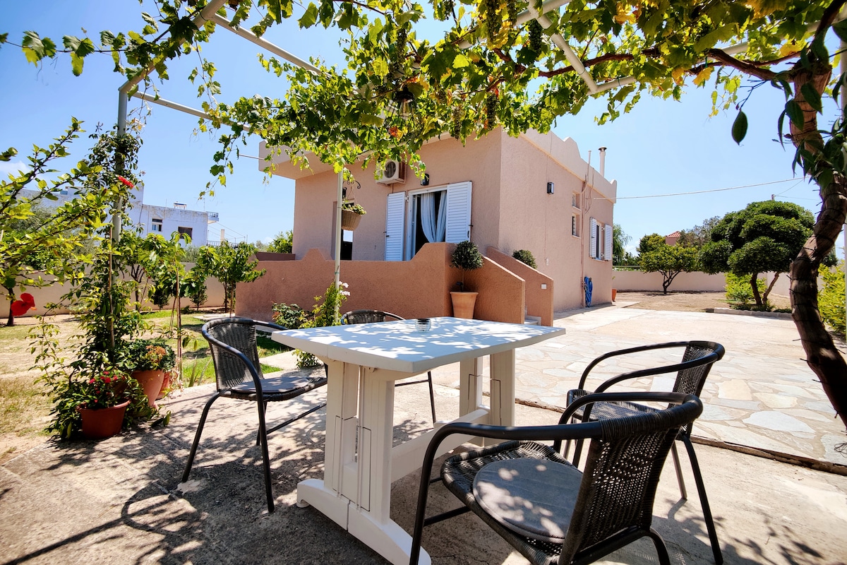 A shaded outdoor seating area is presented, featuring a white table surrounded by four black chairs. The background reveals a detached house with large windows and a well-kept garden, abundant with greenery and citrus trees, all under a clear blue sky.