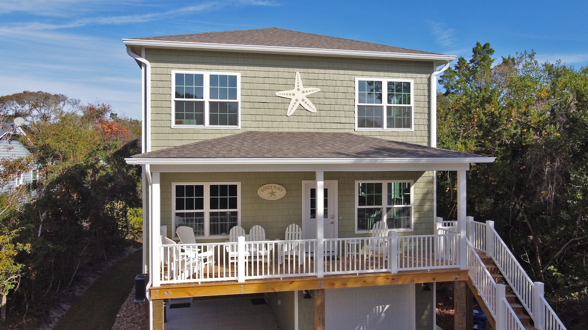 A two-story home on stilts is presented, featuring a welcoming front porch adorned with white rocking chairs. The exterior showcases a light green color with a starfish decoration above the entrance, and four large windows provide natural light.