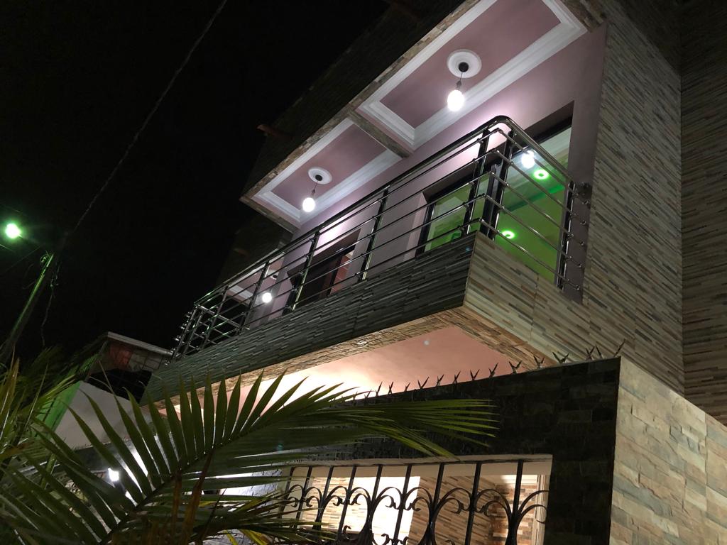 The exterior of the building is viewed at night, showcasing a modern design with a balcony and large windows. Bright lights illuminate the façade, highlighting a mix of textured stone and painted surfaces. Lush greenery is visible in the foreground, adding to the welcoming appearance.