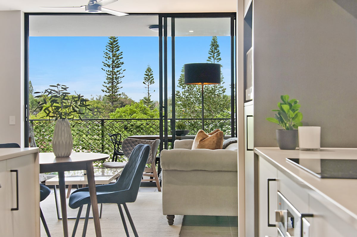An open living area is highlighted by large glass doors leading to a balcony, allowing natural light to fill the space. A modern dining table with four chairs and a cozy sofa are visible. Green plants are placed on tables, enhancing the contemporary design.