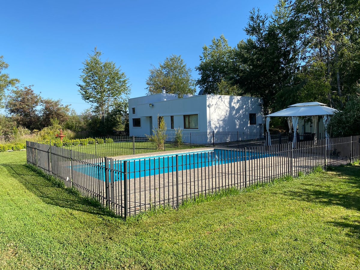 A private swimming pool is enclosed by a wooden fence, surrounded by well-maintained grass and native trees. The white house features multiple windows, allowing natural light to enter. A shaded seating area is visible next to the pool, inviting relaxation in a tranquil environment.