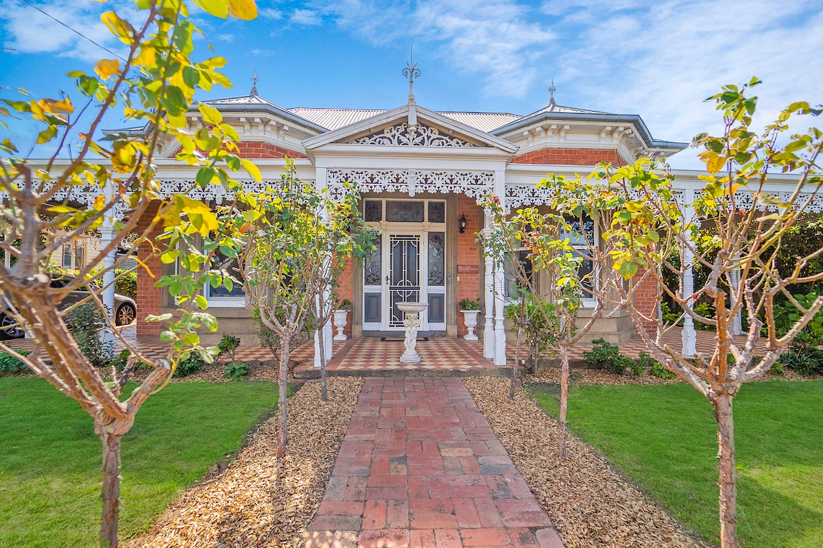The entrance features a charming Victorian façade with intricate metalwork and a tiled path leading to the front porch. Flanking the path are well-tended garden beds with small trees, complemented by lush green grass providing a welcoming approach to the home.