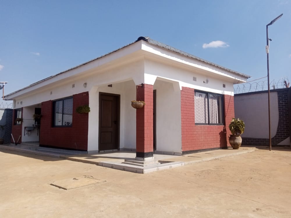 The exterior of the house is presented, showcasing a single-story structure with a combination of red brick and white walls. Large windows allow natural light to enter, while a porch area is visible at the entrance. A potted plant is placed near the door, and the surrounding area is paved.