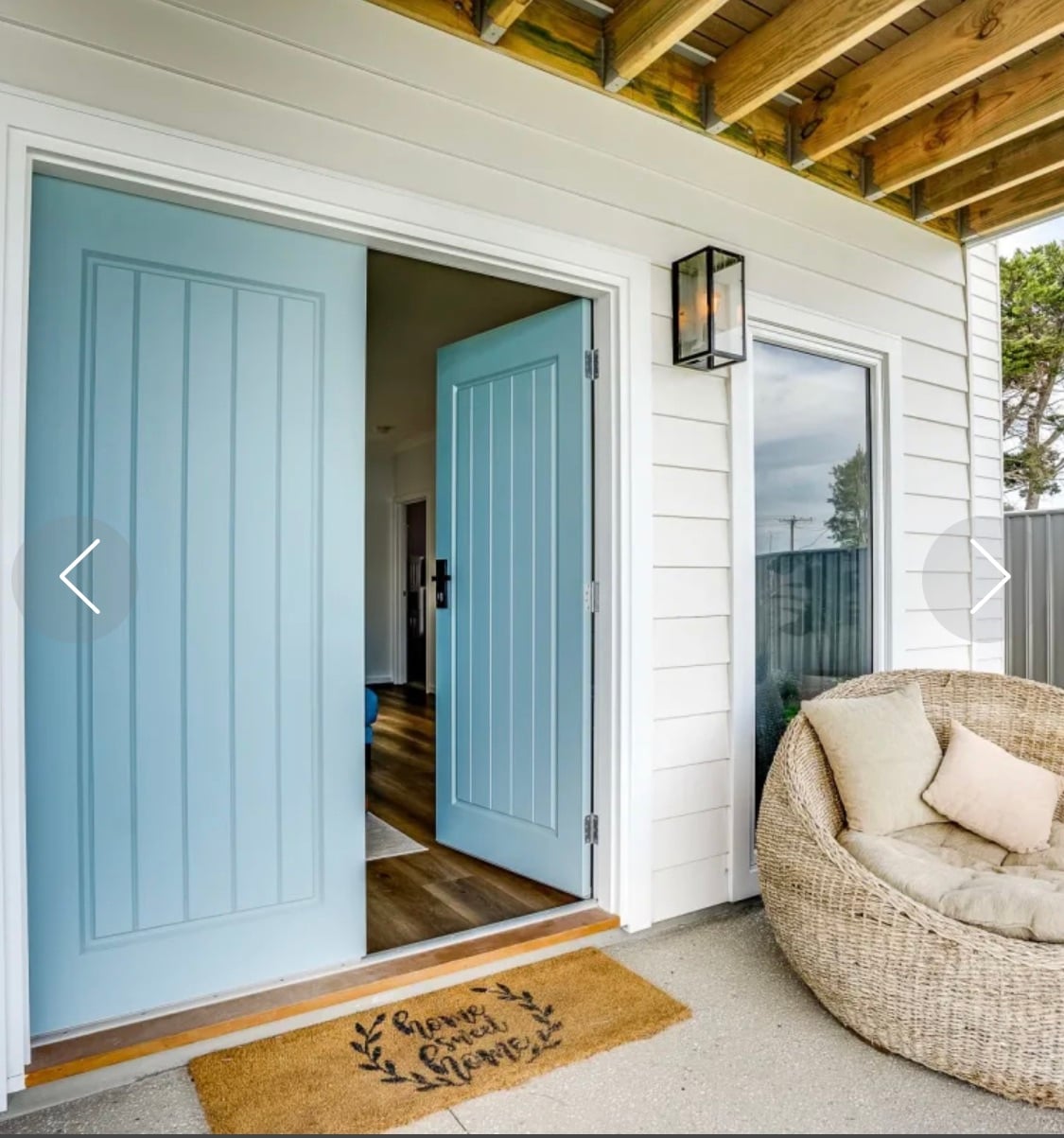 The image shows a welcoming front entrance with two light blue doors that are open wide. A cozy wicker chair is positioned nearby. A doormat with the message 'Home Sweet Home' is placed just outside the entrance, suggesting a friendly and inviting atmosphere.