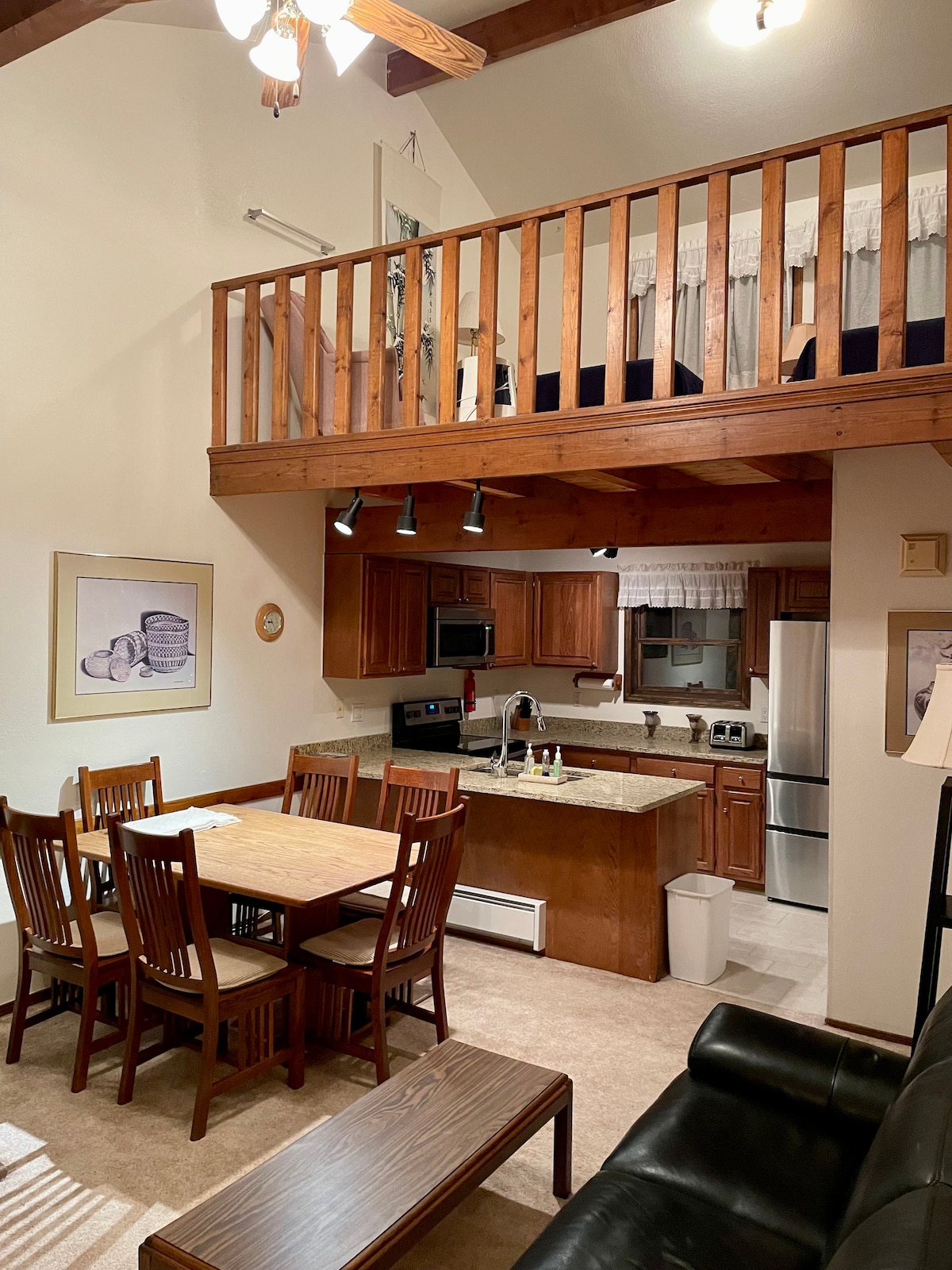 An open living area showcases a dining table surrounded by wooden chairs, complemented by a kitchen featuring wood cabinetry and stainless-steel appliances. A lofted sleeping area is visible above, accessed by a wooden railing, with light fixtures illuminating the space.